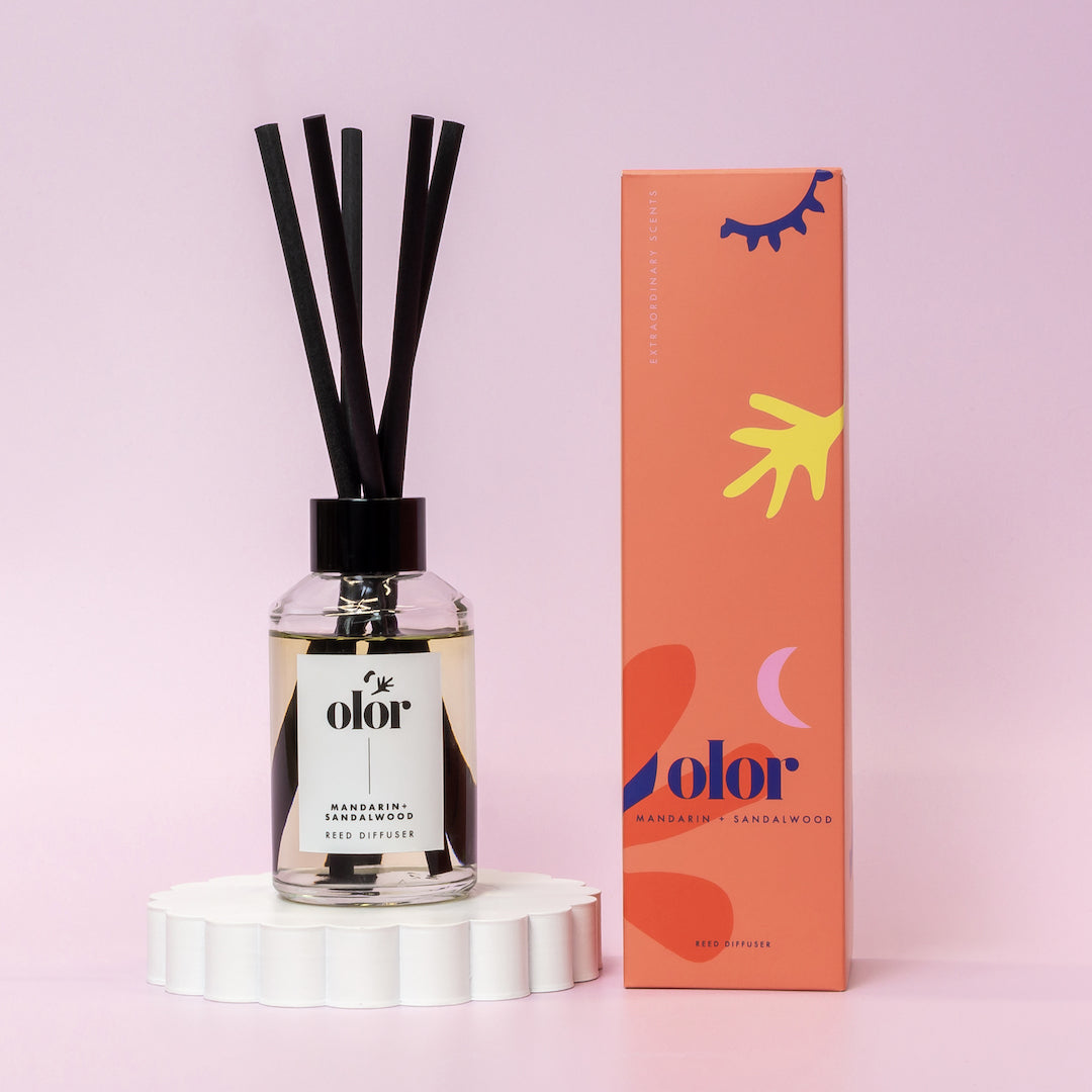 Olor Mandarin & Sandalwood reed diffuser in clear glass bottle with black reeds, displayed on a white stand next to its