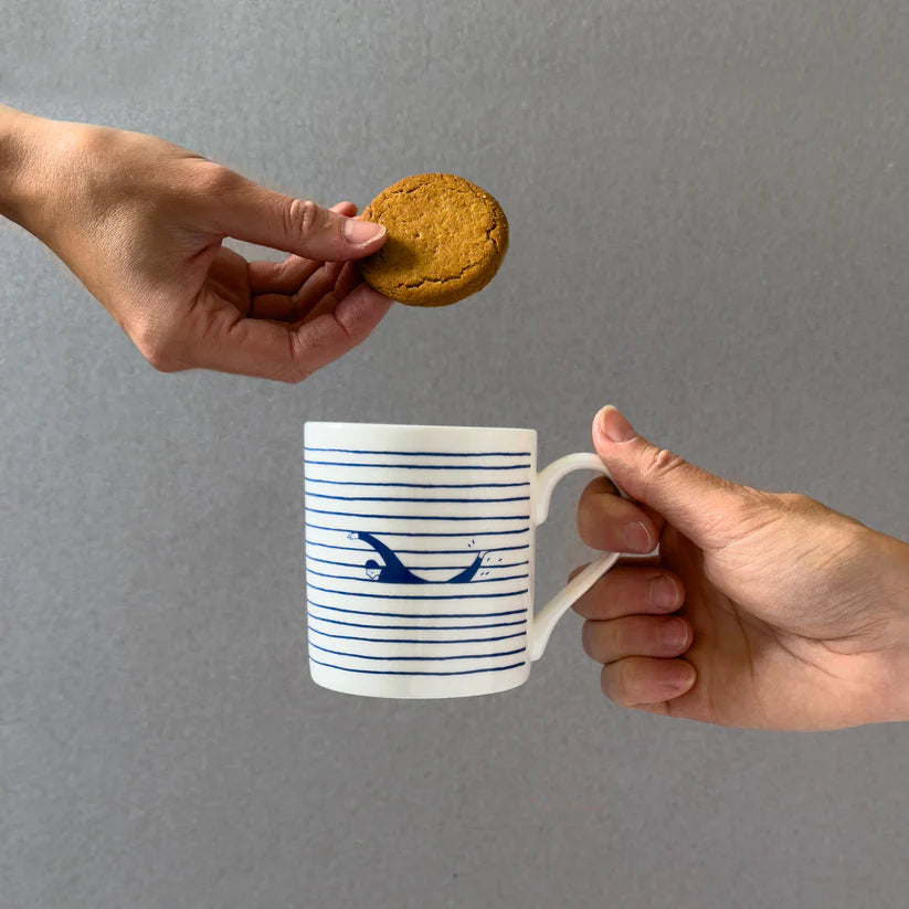 White mug with blue horizontal stripes and a swimmer design, a ginger biscuit being offered to it.
