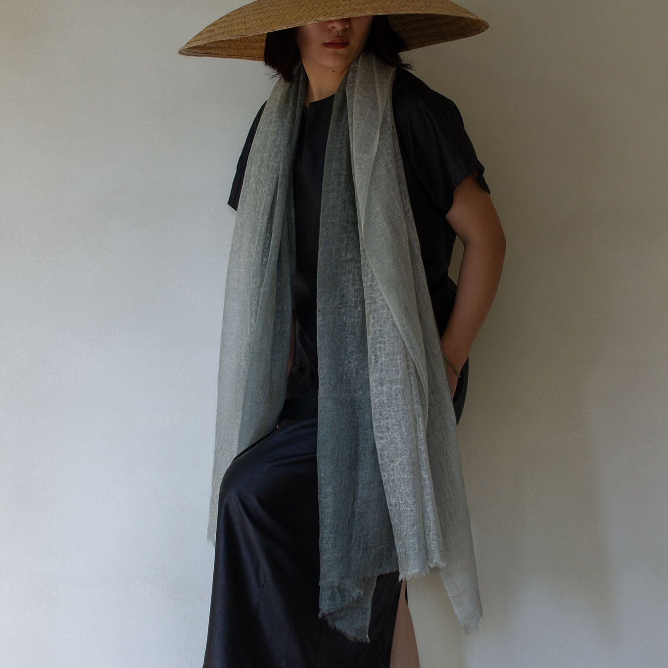 A person wearing a large straw hat and a long, sage green dip-dyed cashmere scarf with a black top and pants against a neutral background.