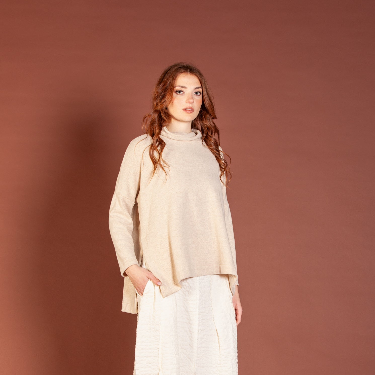 Woman wearing a beige sweater and white pants against a brown background
