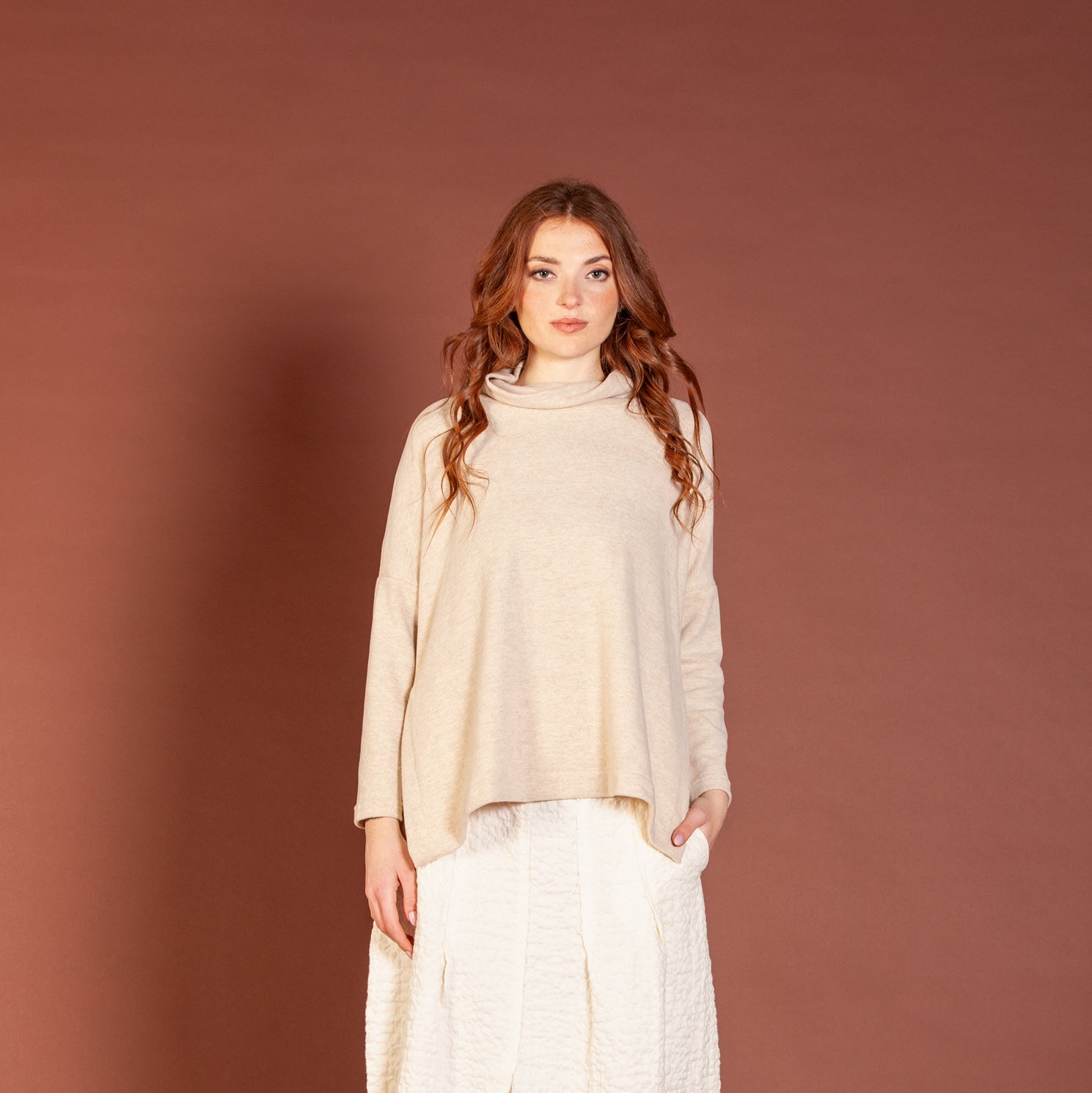 Woman wearing a beige sweater and white pants against a brown background