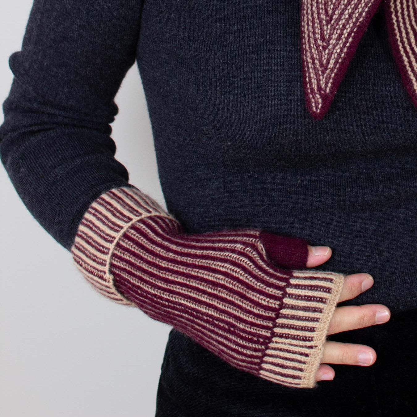 A close-up of a person wearing a dark navy ribbed sweater and burgundy and beige striped fingerless knit gloves, with a matching burgundy and beige striped scarf draped over the shoulders.