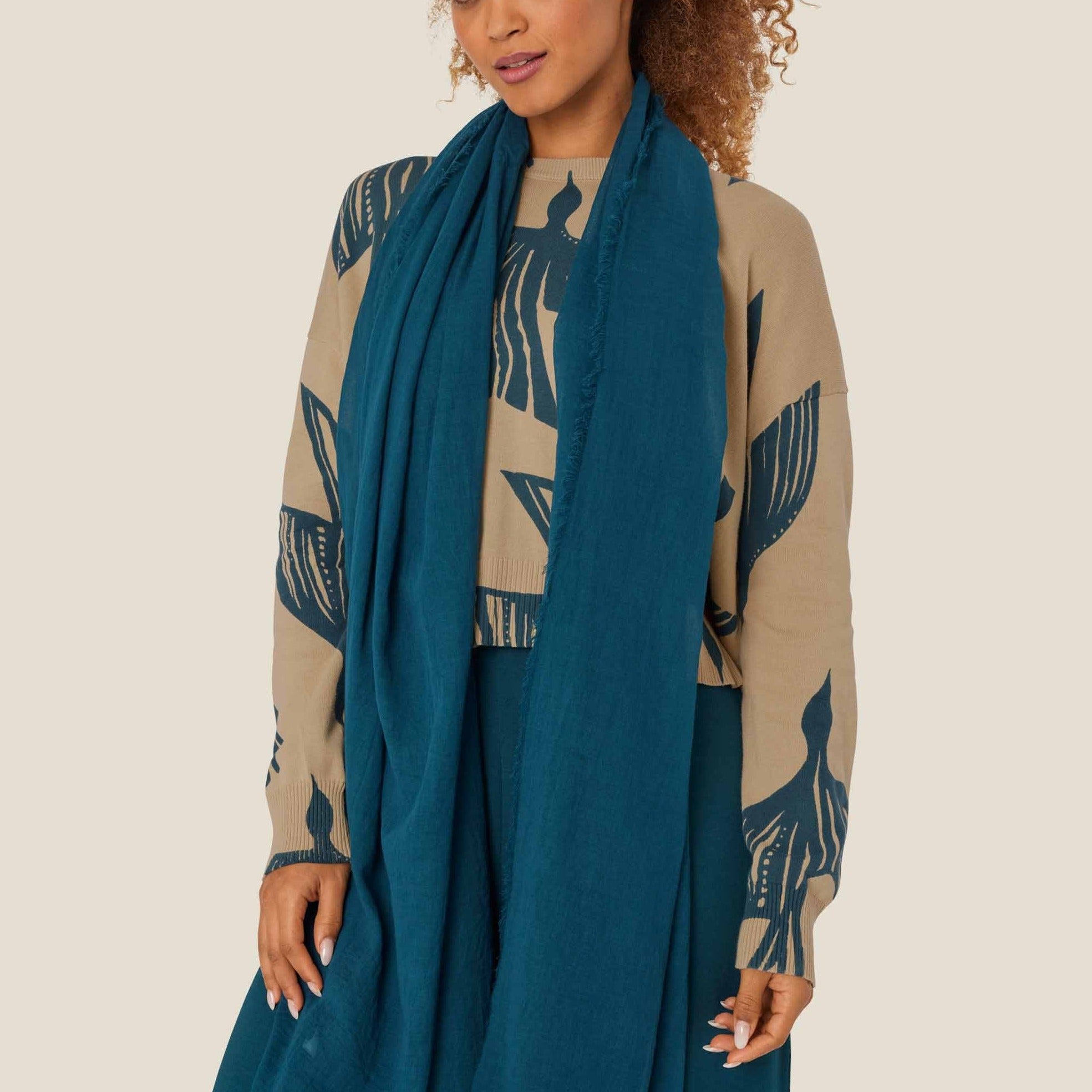 Teal rectangular scarf with frayed edges, draped around a person wearing a beige sweater with dark teal bird print.