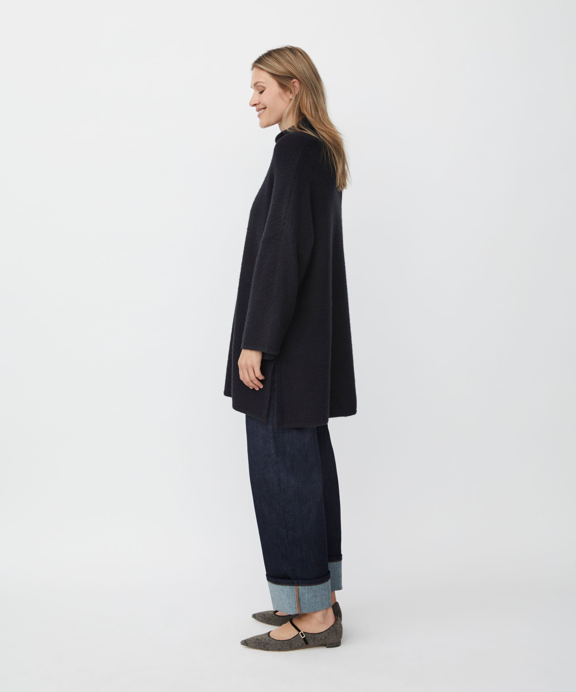 Woman wearing a black tunic and dark jeans standing against a white background