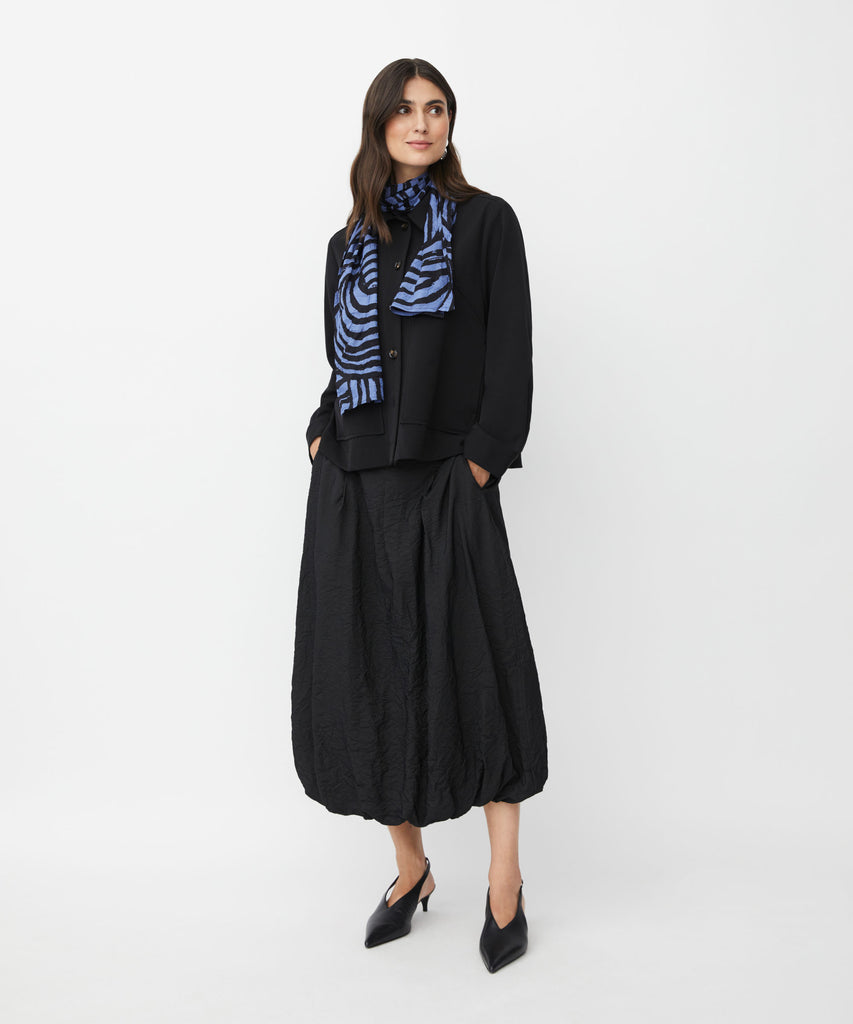 Black cropped button-front jacket, worn with a blue and black patterned scarf and a textured black midi skirt.