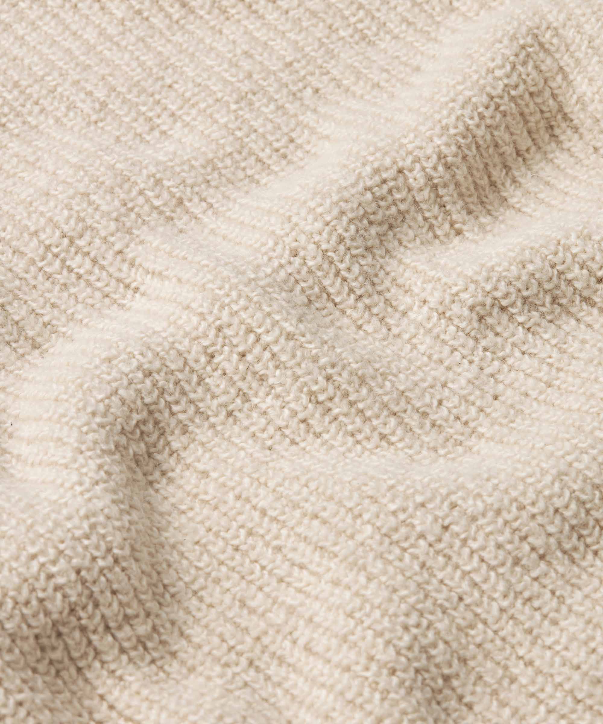 Close-up of a cream-colored knit fabric with a textured waffle or ribbed pattern, showing soft folds and shadows.