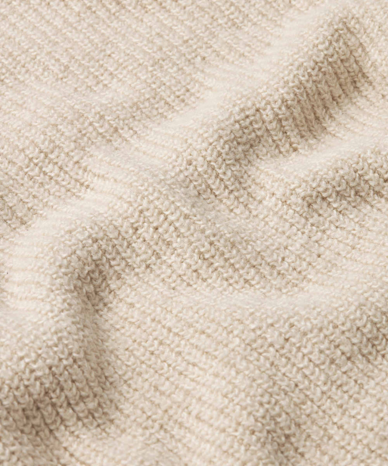 Close-up of a cream-colored knit fabric with a textured waffle or ribbed pattern, showing soft folds and shadows.