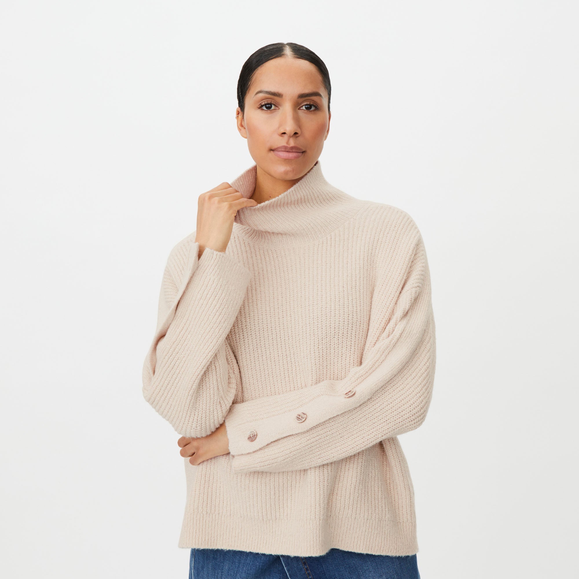 A woman wearing a light beige ribbed knit turtleneck sweater with wide balloon sleeves and decorative buttons on the cuffs, paired with blue jeans, against a plain white studio background.