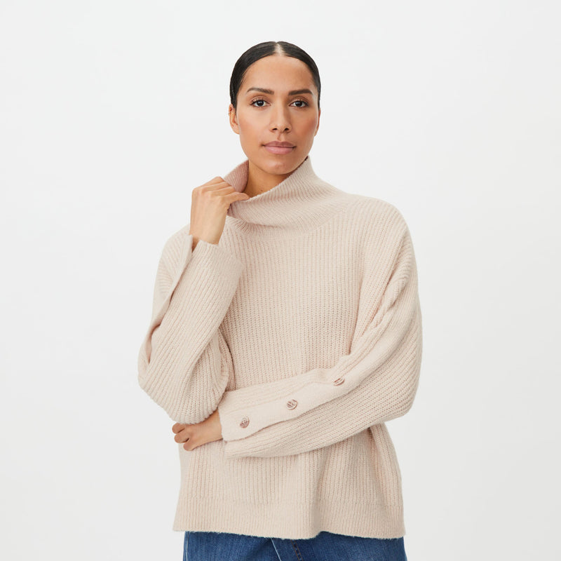 A woman wearing a light beige ribbed knit turtleneck sweater with wide balloon sleeves and decorative buttons on the cuffs, paired with blue jeans, against a plain white studio background.