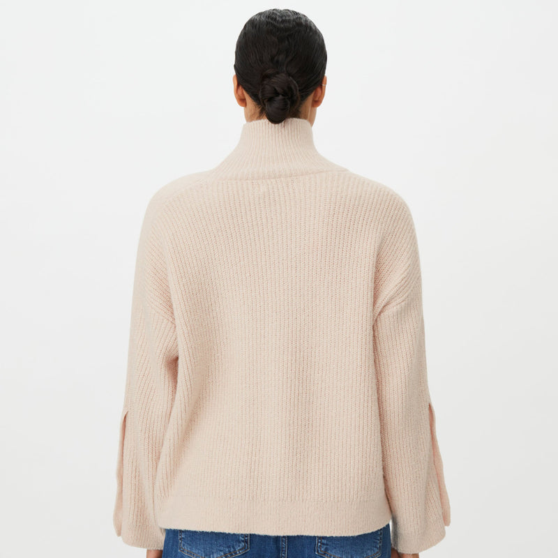 A model viewed from the back wearing a light beige ribbed knit mock-neck sweater and blue jeans against a plain white background.