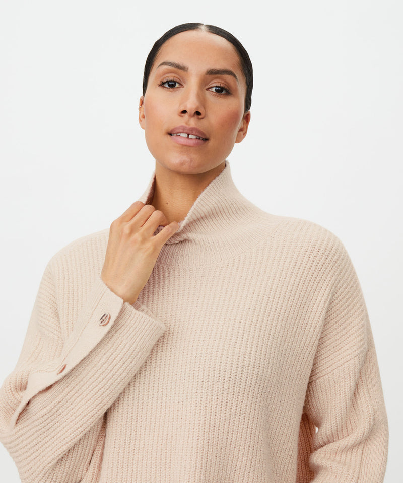 A woman wearing a light beige ribbed knit mock-neck sweater, posing against a clean white studio background.