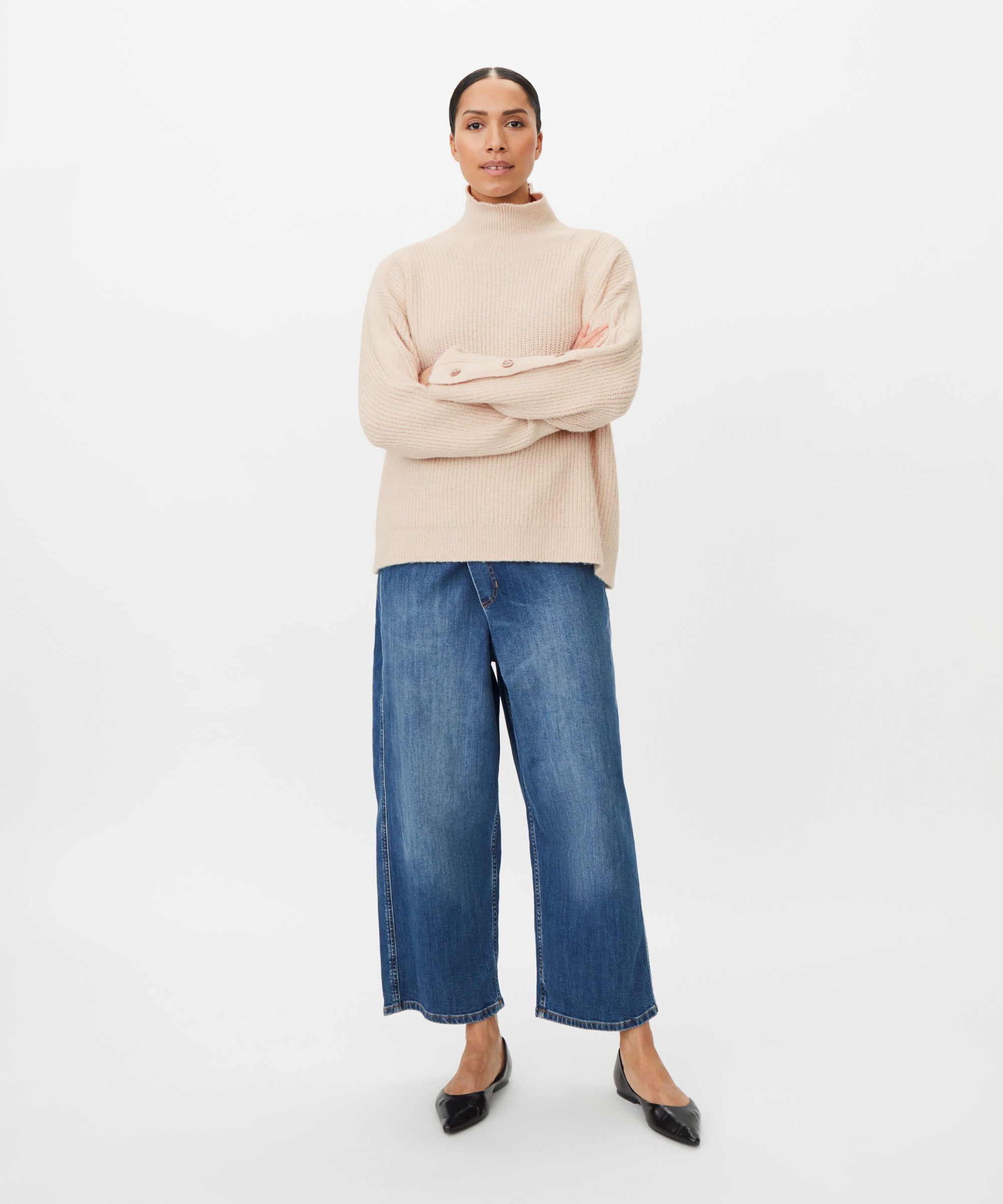 A model stands against a plain white studio background wearing a light beige turtleneck sweater, wide-leg blue jeans, and black pointed flats.