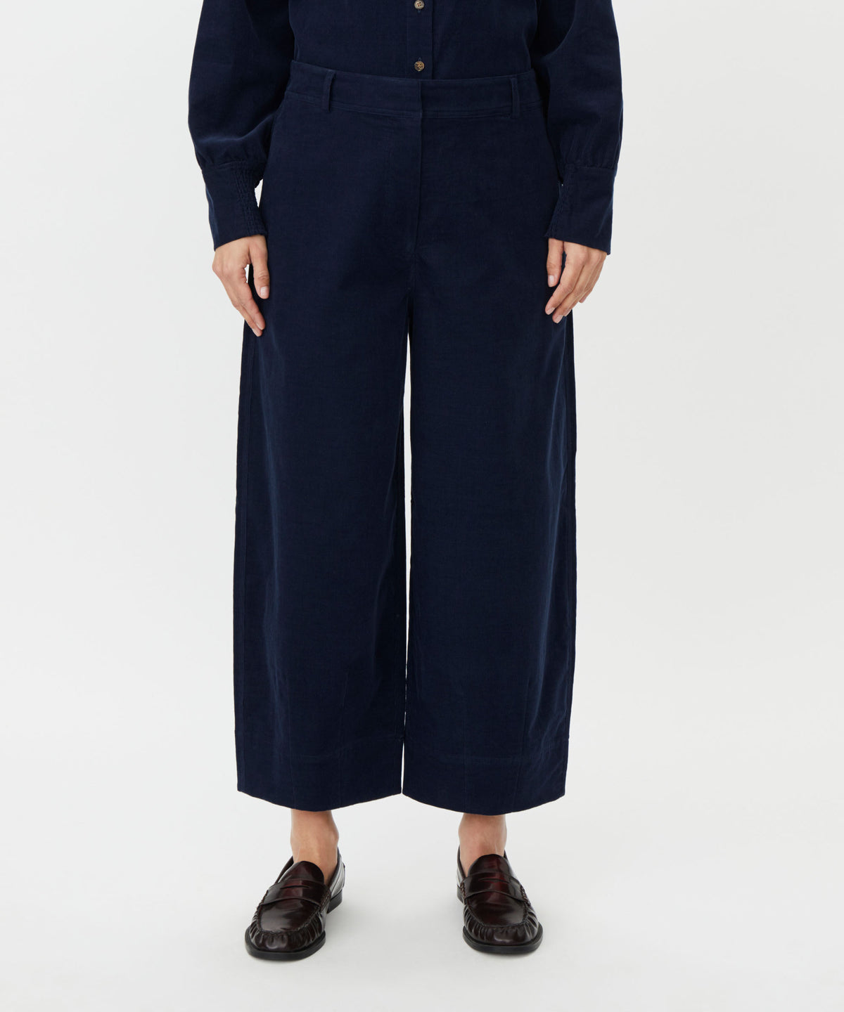 A model wearing dark navy blue wide-leg cropped trousers with a matching long-sleeve button-up top and dark brown leather loafers, shown from the waist down on a light gray background.