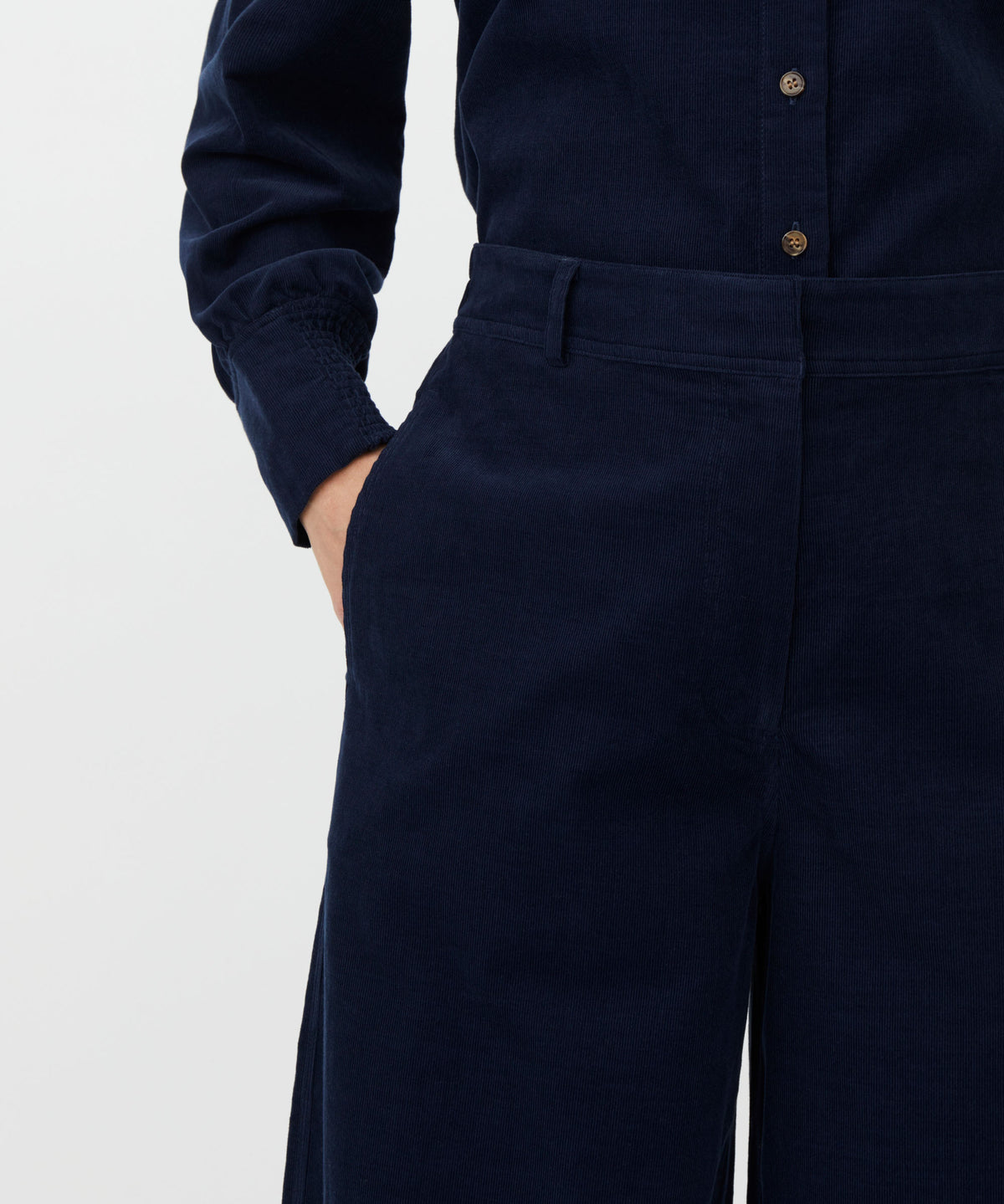 Close-up of a person wearing dark navy corduroy trousers with a hand in the pocket, paired with a matching navy long-sleeve button-up shirt, against a light gray background.