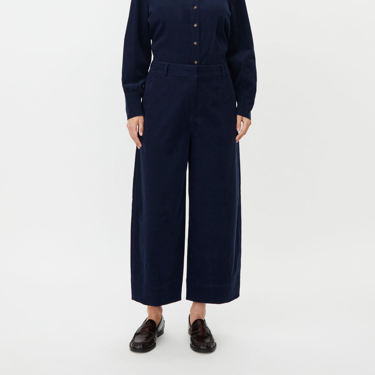 A woman stands against a plain light gray studio background wearing a dark navy long-sleeve button-front top and matching wide-leg trousers, paired with dark loafers.