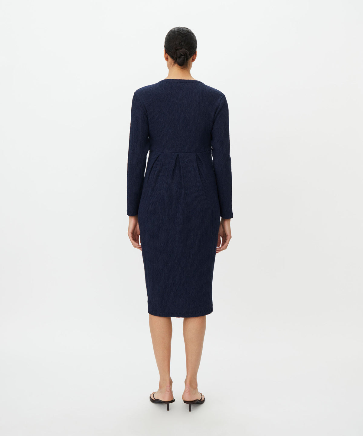 A woman viewed from the back wearing a dark navy knee-length dress with long sleeves and a gathered waist seam, paired with black flat sandals, against a plain white studio background.