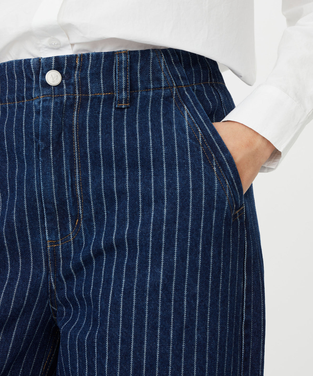 Close-up of a person wearing dark blue pinstriped denim pants with a white button and a white shirt tucked in, against a light gray background.