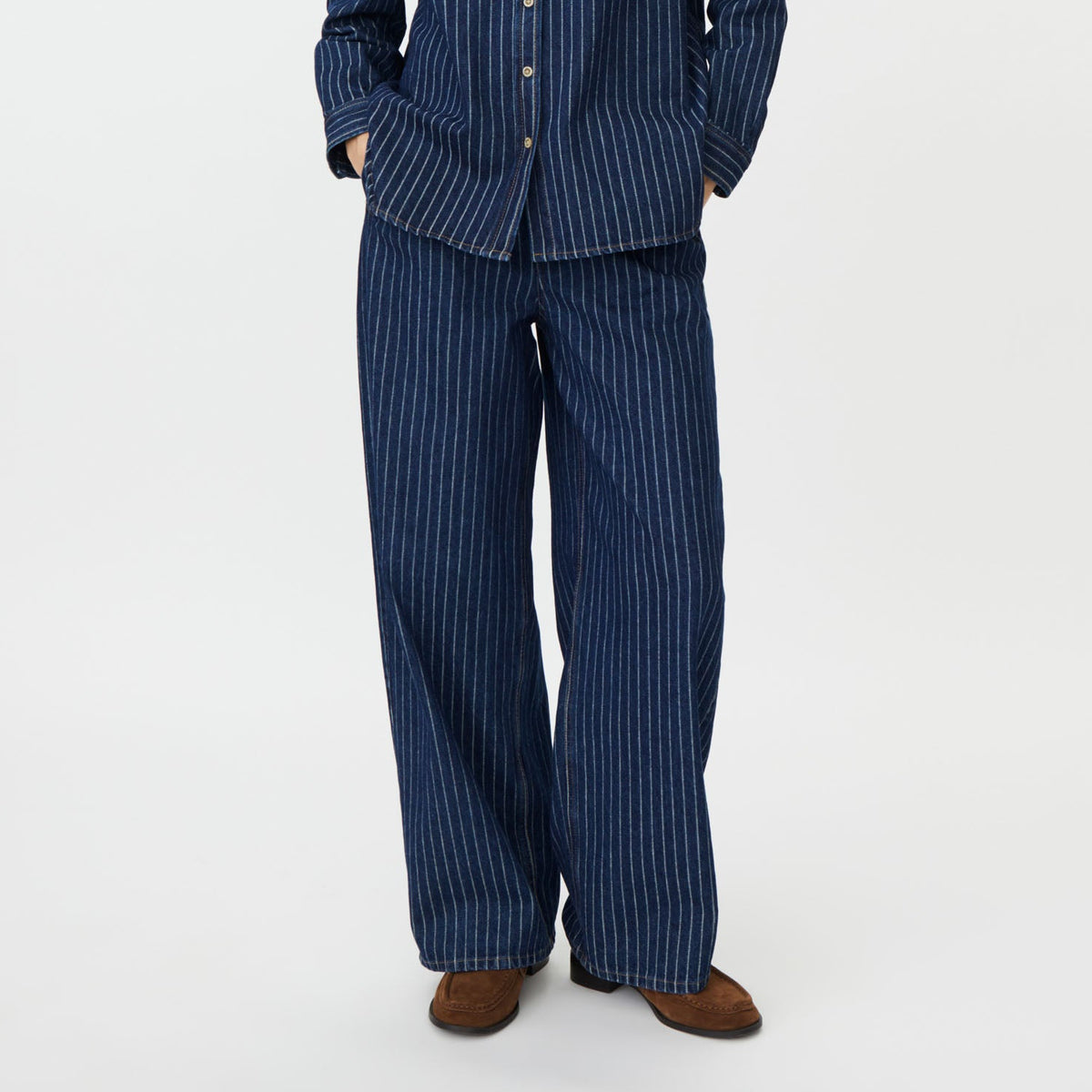 A woman modeling a matching navy blue pinstriped pajama set, featuring a long-sleeve button-up shirt and wide-leg trousers, against a plain white studio background.