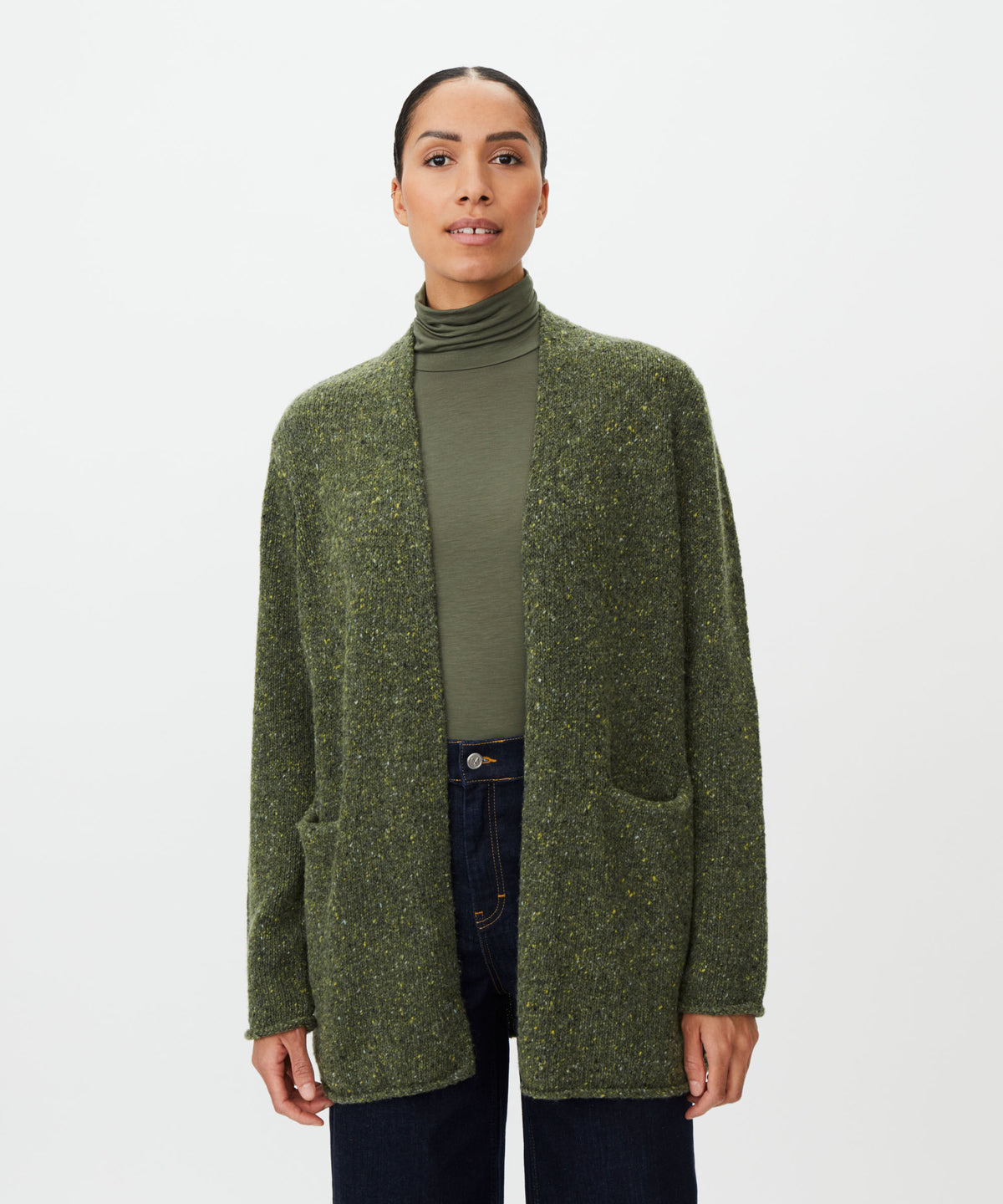 A woman modeling a textured olive green open-front cardigan over a matching olive green turtleneck and dark blue jeans, shot against a plain light gray studio background.