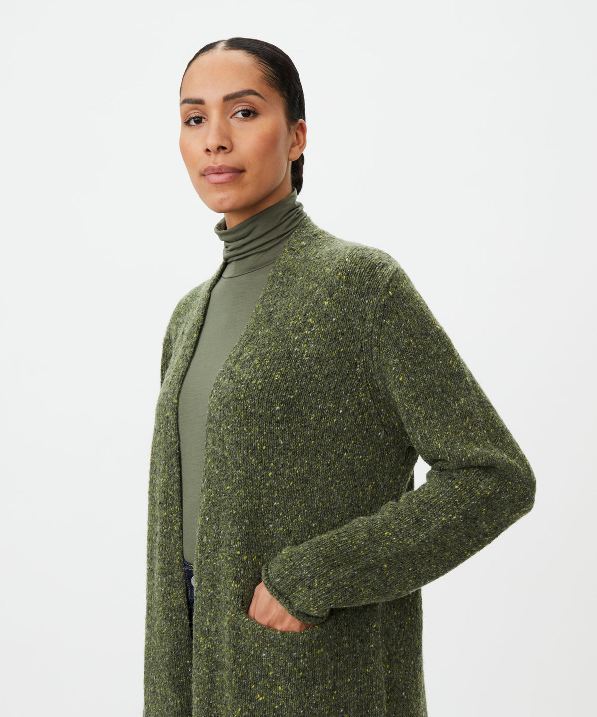 A woman wearing a textured olive green open-front cardigan over a matching green turtleneck sweater, photographed against a clean light gray studio background.