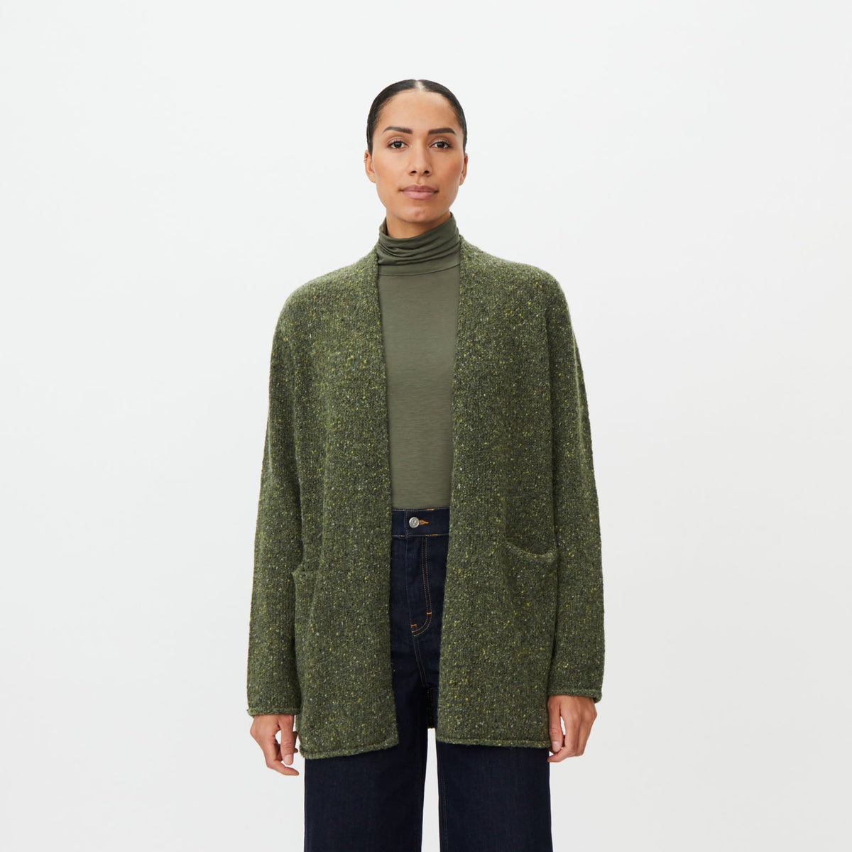 A model stands against a light gray studio background wearing a textured olive green open-front cardigan over a matching olive green turtleneck, dark wide-leg jeans, and brown leather shoes.