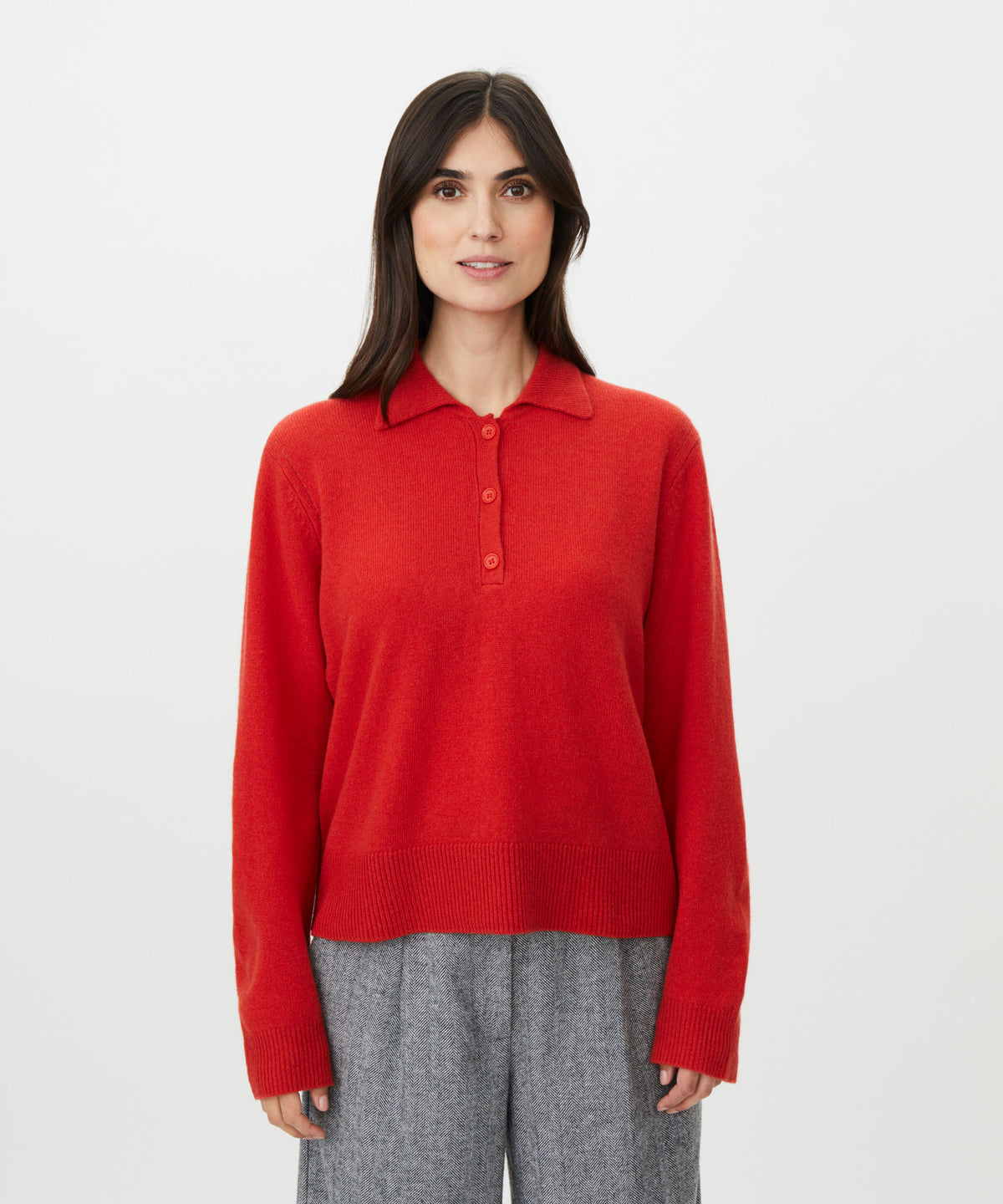 A woman modeling a bright red knit polo sweater with a three-button placket, paired with gray trousers, against a plain light gray studio background.