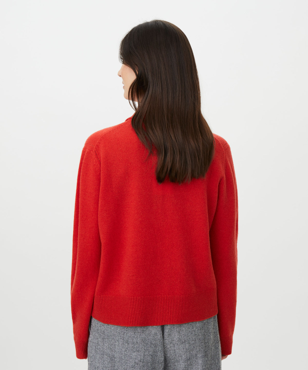 A woman with long dark brown hair is shown from the back wearing a bright red knit sweater and gray trousers against a plain light gray studio background.