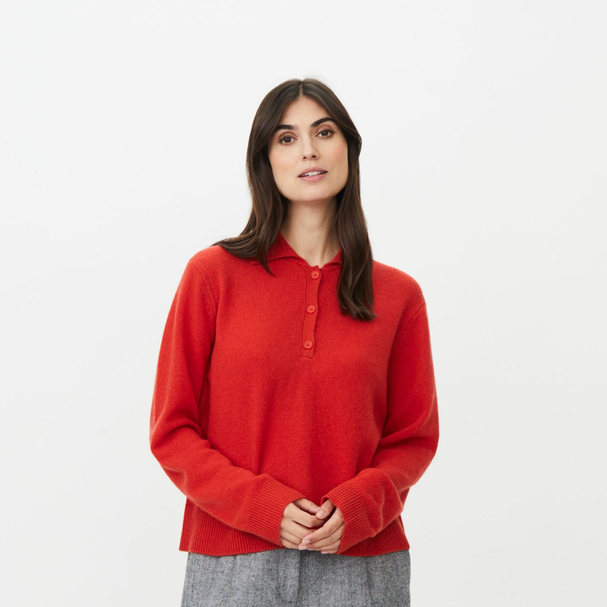 A woman modeling a red long-sleeve polo-style knit top with a partial button placket, paired with wide-leg gray trousers and dark shoes, against a plain light gray studio background.