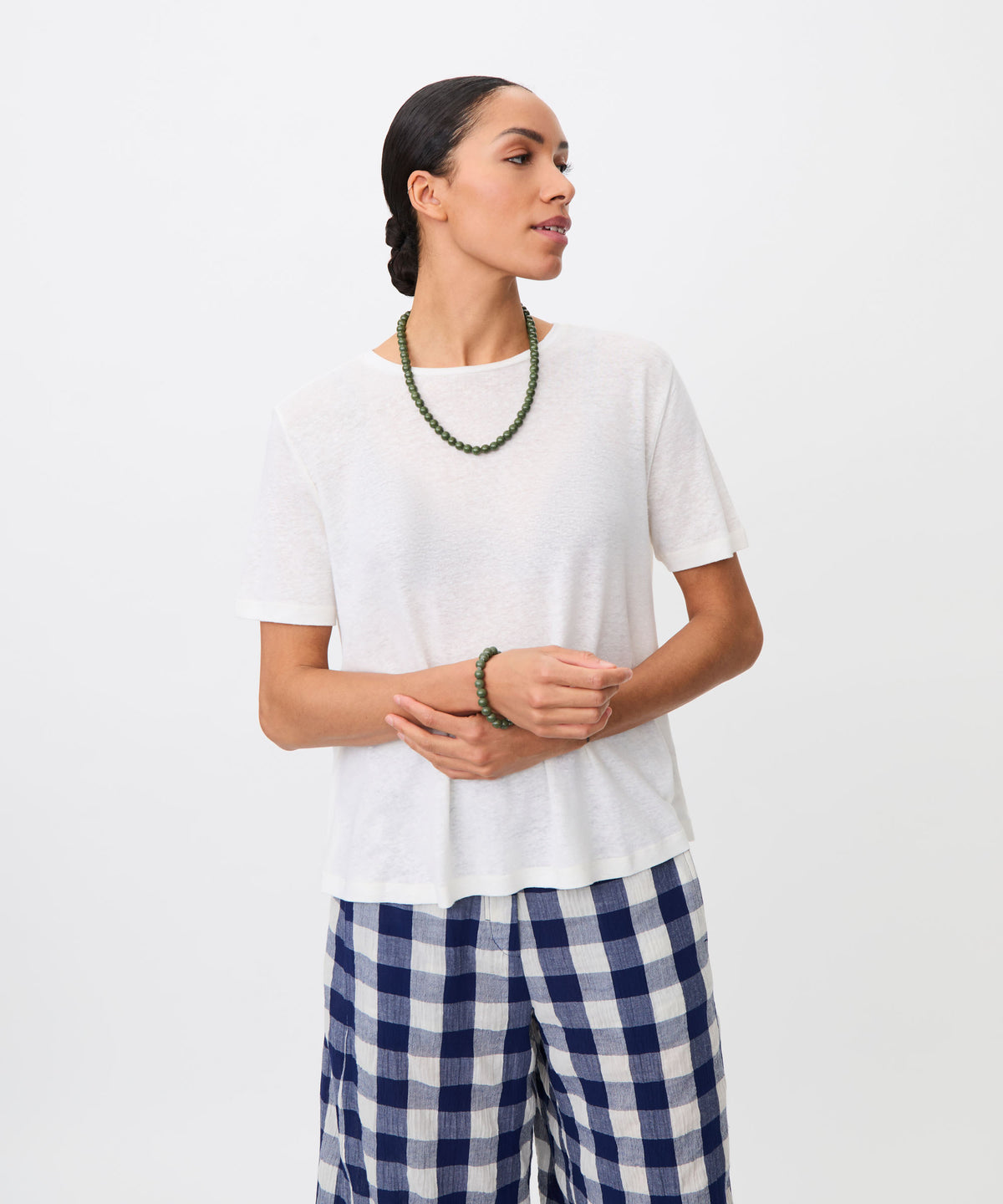 A woman modeling a white short-sleeve crewneck t-shirt, paired with blue and white checkered pants, a green beaded necklace, and a matching green beaded bracelet, against a plain white background.