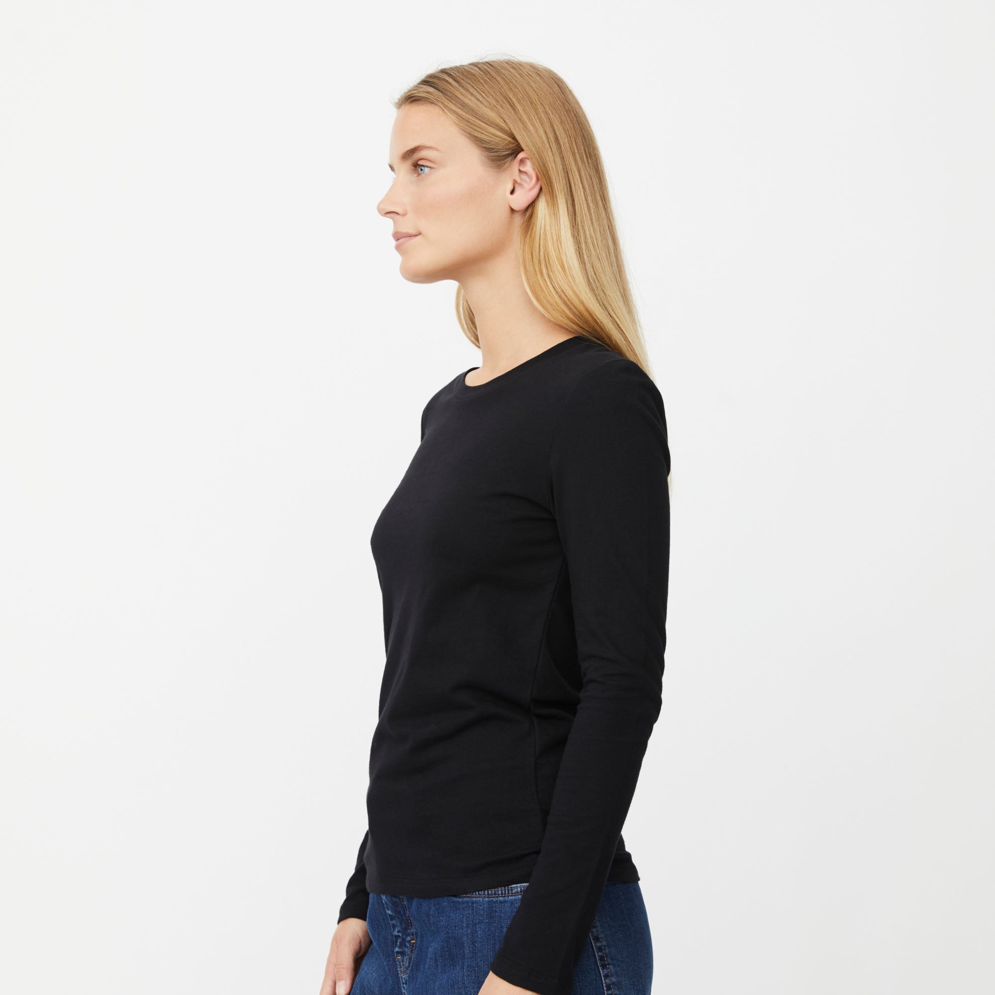 A woman in profile wearing a black long-sleeve crewneck top and blue jeans, photographed against a clean white studio background.