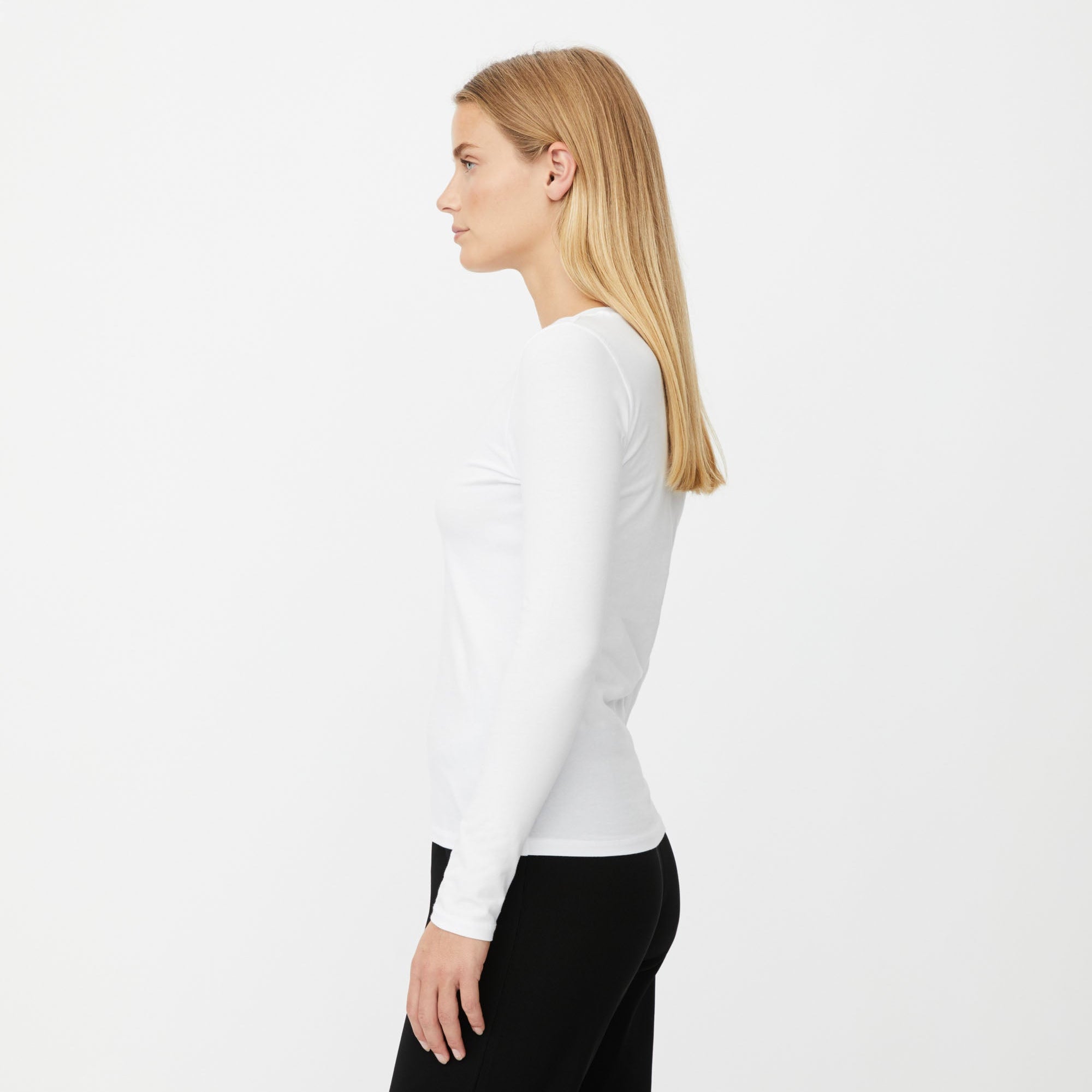 A woman in profile wearing a fitted white long-sleeve top and black pants, photographed against a clean white studio background.