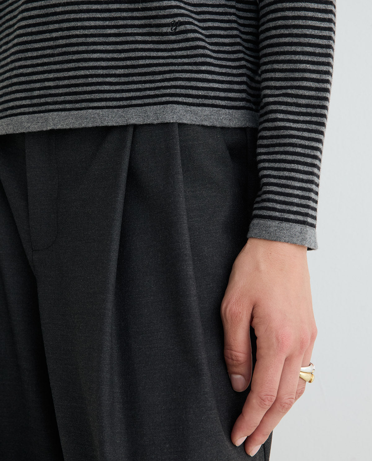 Close-up of a person wearing a dark gray pleated wool skirt or trousers and a black and gray striped knit top, with a gold ring on one finger against a light gray background.
