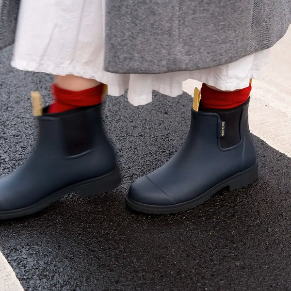 Person wearing black rain boots with red socks on a gray pavement.