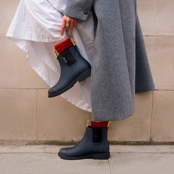 Person wearing black rain boots with red socks and a gray coat against a beige wall.