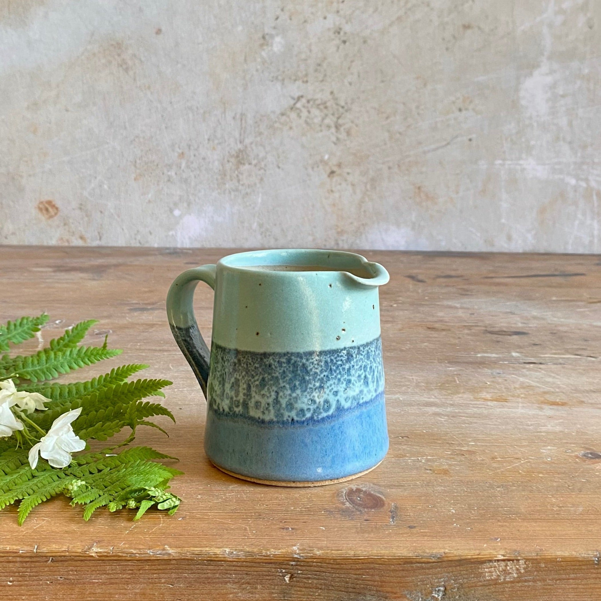Small, handcrafted ceramic creamer or milk jug.  Light teal top transitioning to darker blue and speckled glaze at the base.