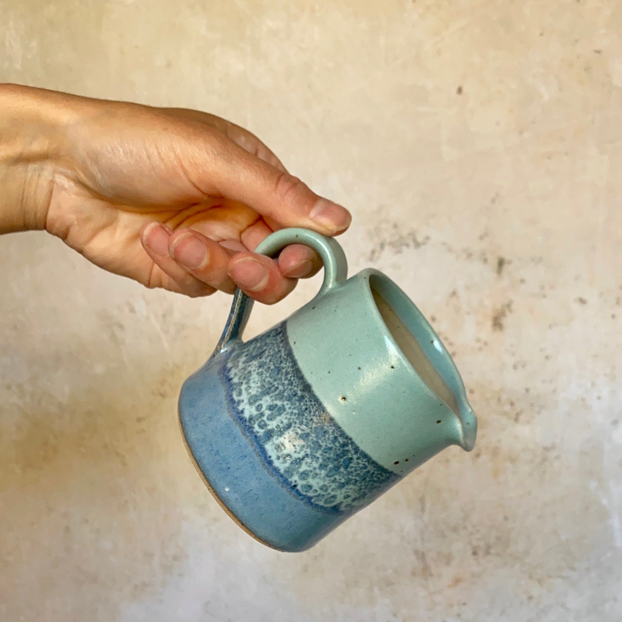 Hand holding a small, speckled, blue and teal ceramic creamer.