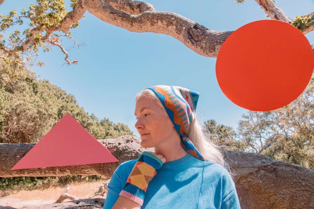 A woman outdoors under a tree wearing a bright blue shirt and a colorful patterned headscarf, with a large red circular graphic overlay on the right and a pink triangular graphic overlay on the left.