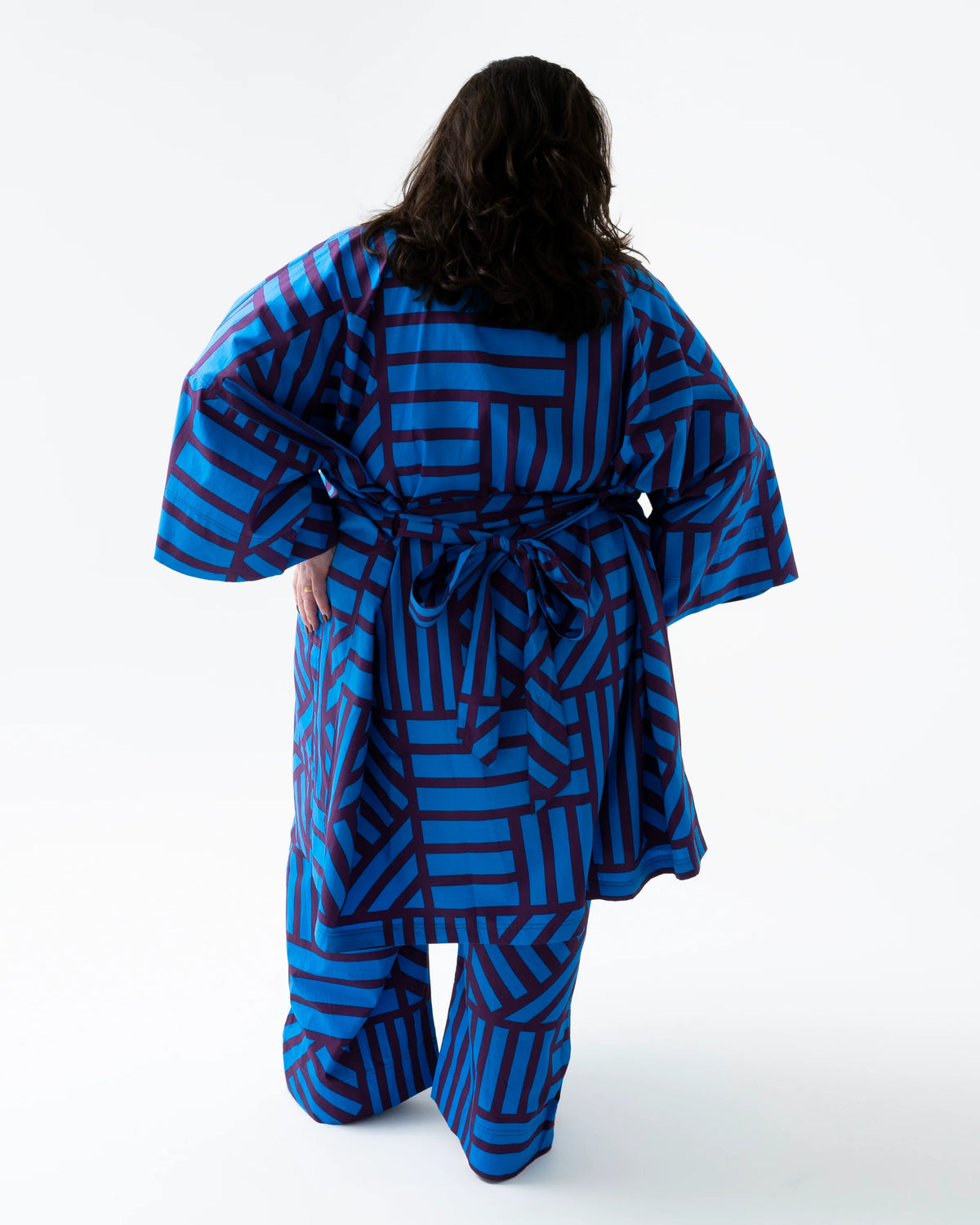 A person shown from the back wearing a loose-fitting robe and matching pants with a bold blue and dark red geometric stripe pattern, photographed against a clean white studio background.