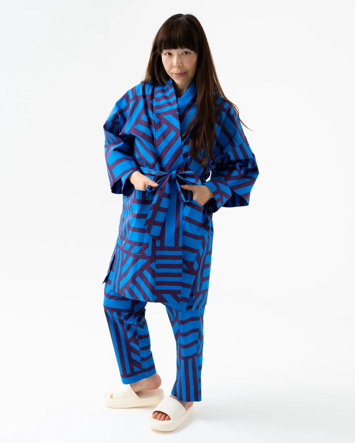 A woman modeling a matching two-piece pajama set featuring a bold geometric pattern in royal blue and dark navy, styled with white slide sandals against a plain white studio background.