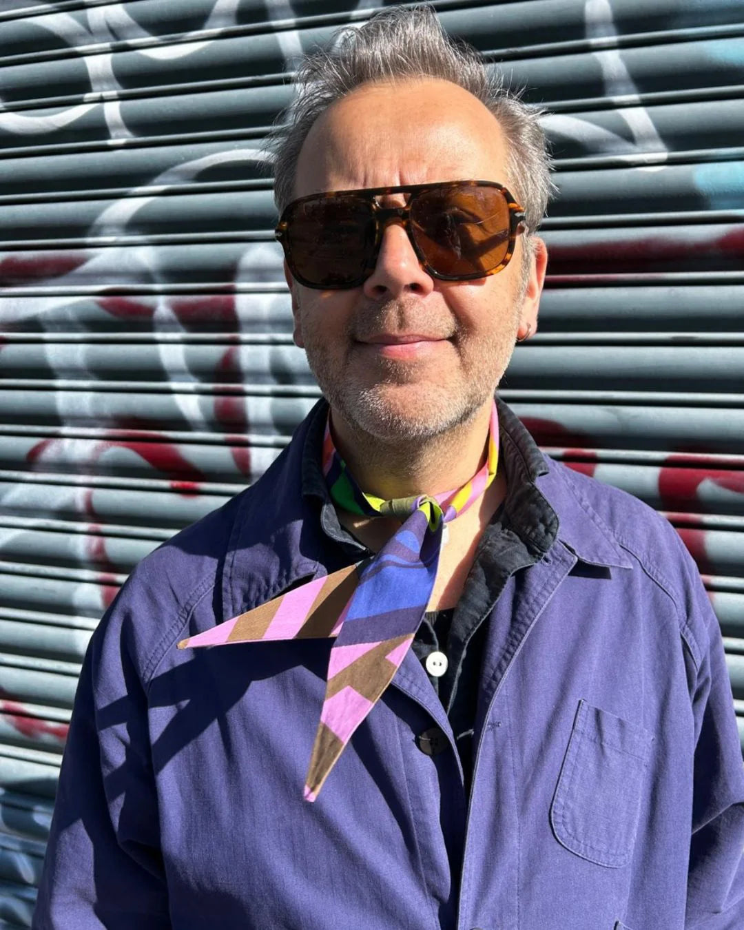 A man wearing large brown-tinted sunglasses, a purple overshirt, and a colorful geometric neck scarf stands in front of a graffiti-covered metal shutter.