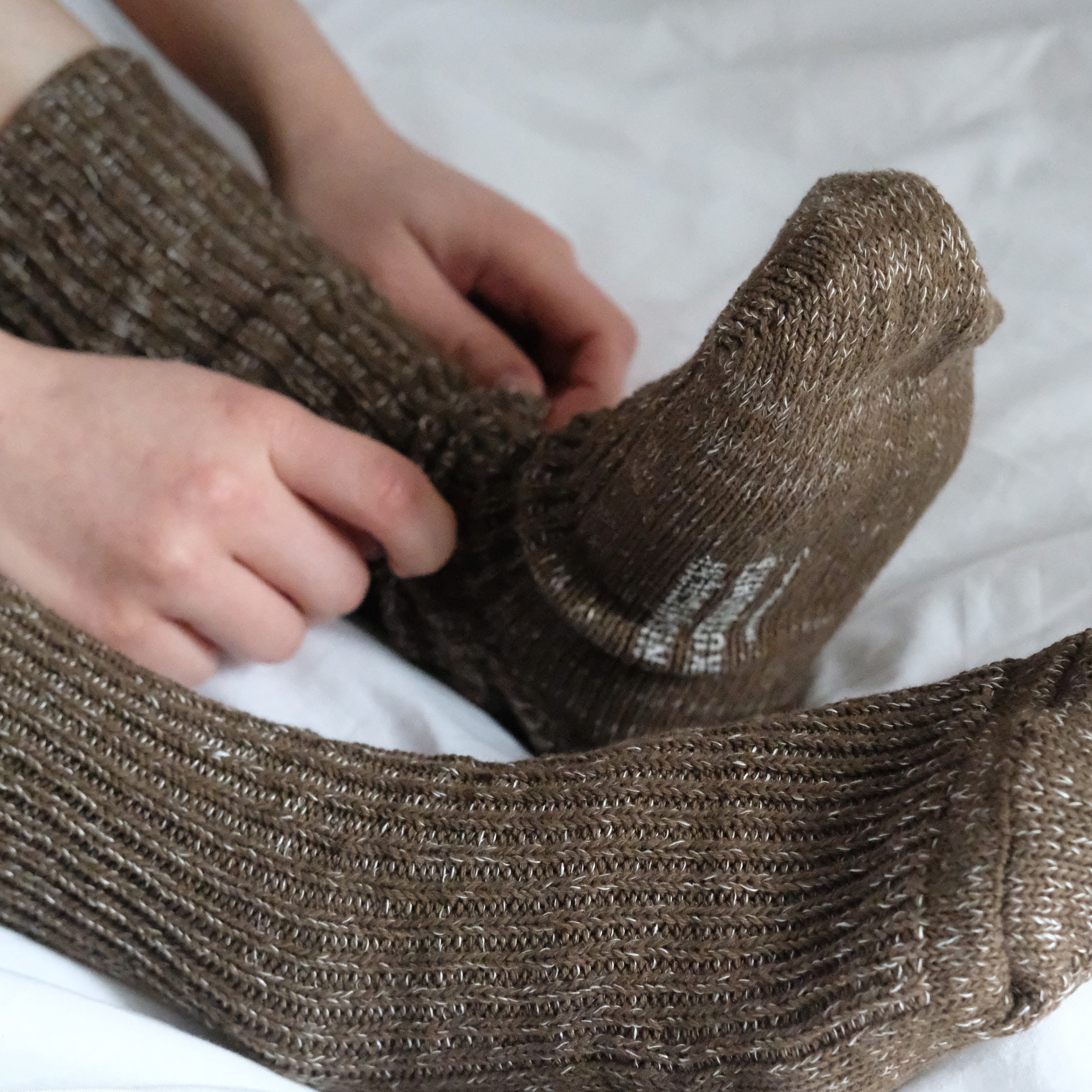 Pair of brown ribbed knit socks with subtle flecks of lighter brown and white.  A person's hands are pulling one sock up.