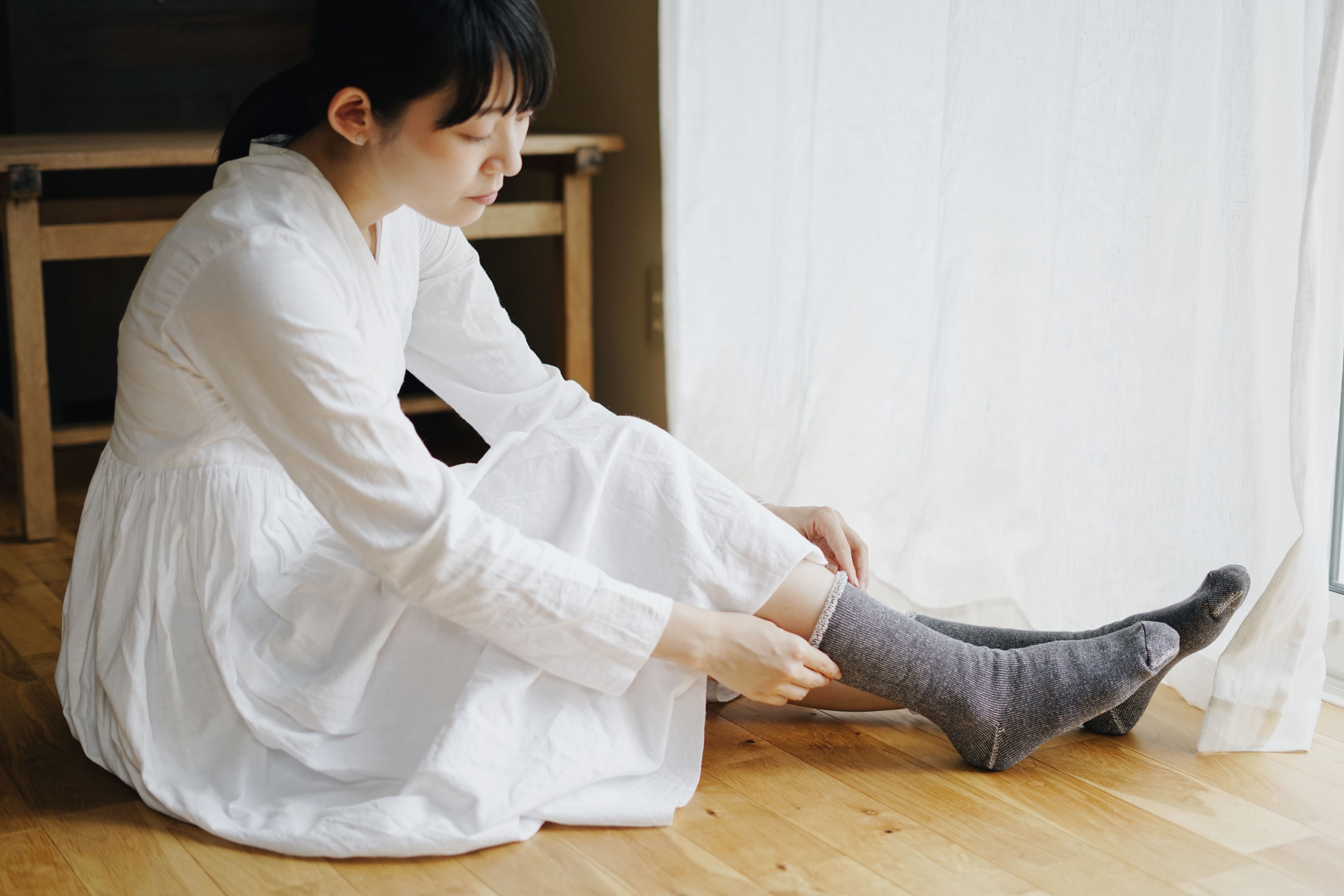 A person in a white dress sits on a hardwood floor, putting on a pair of dark gray, speckled socks with a lighter gray cuff.