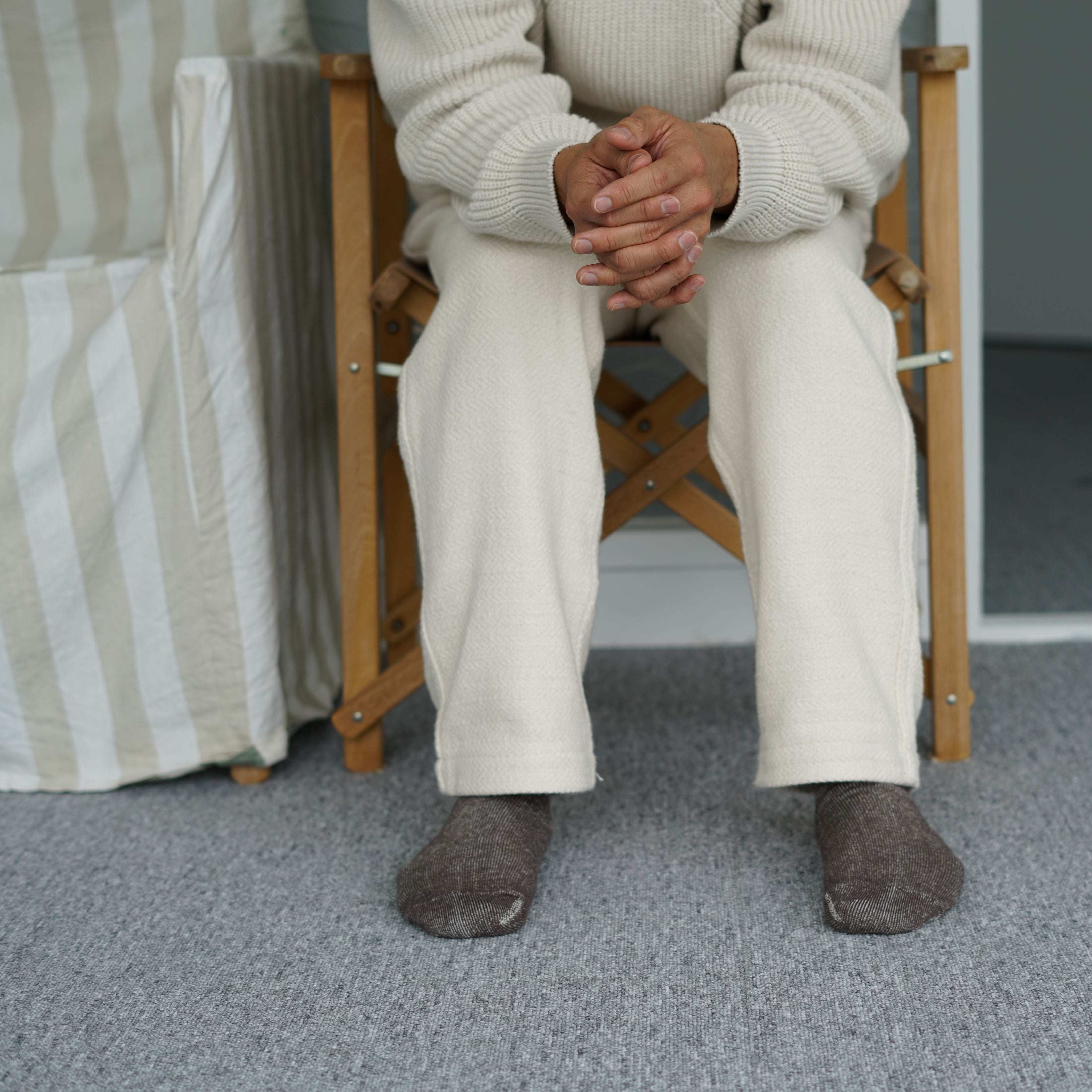 Person seated in a wooden chair wearing off-white sweater and pants, and dark brown socks.