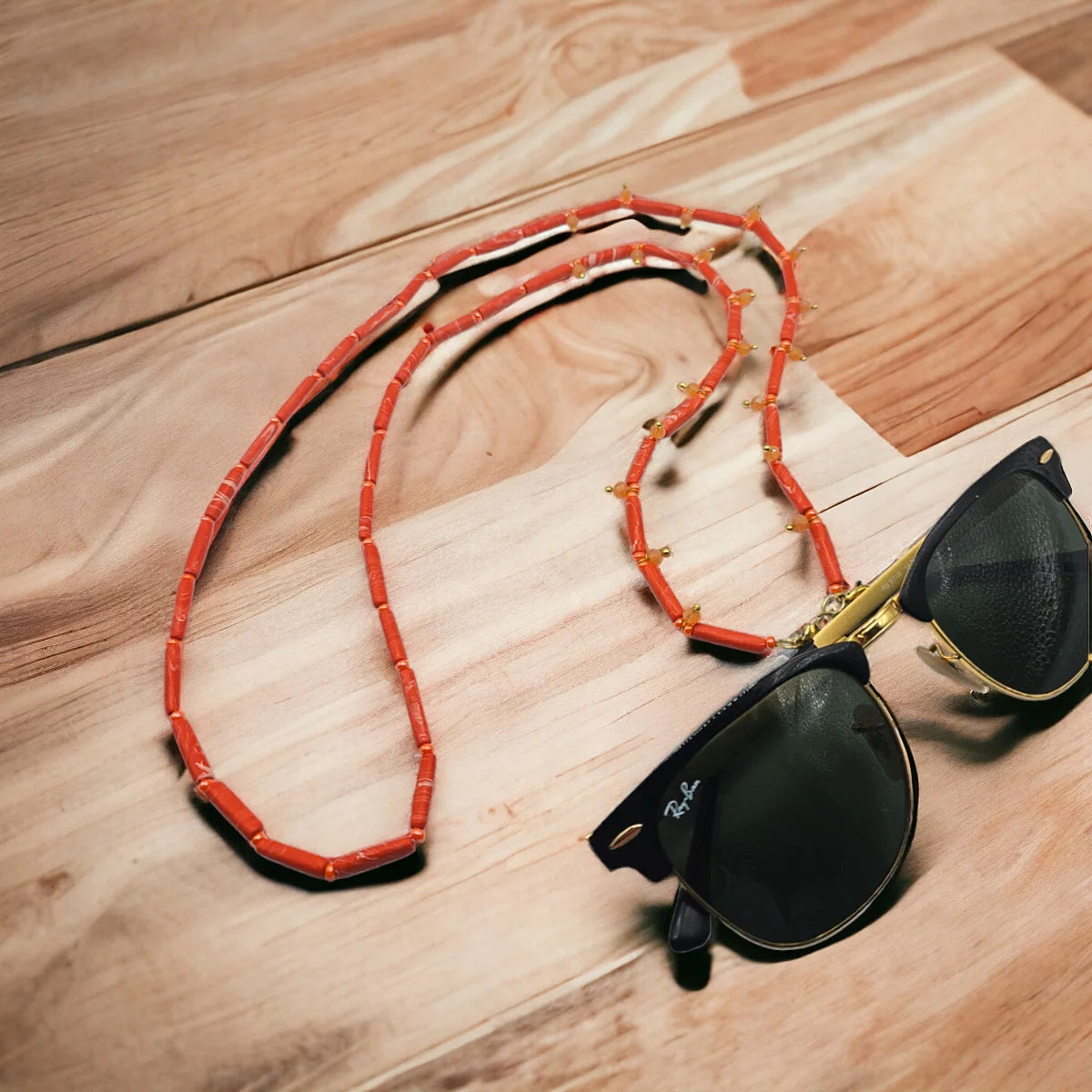 A coral-red beaded necklace with small gold-toned spacer beads, styled on a light wood surface next to black sunglasses with gold frames.
