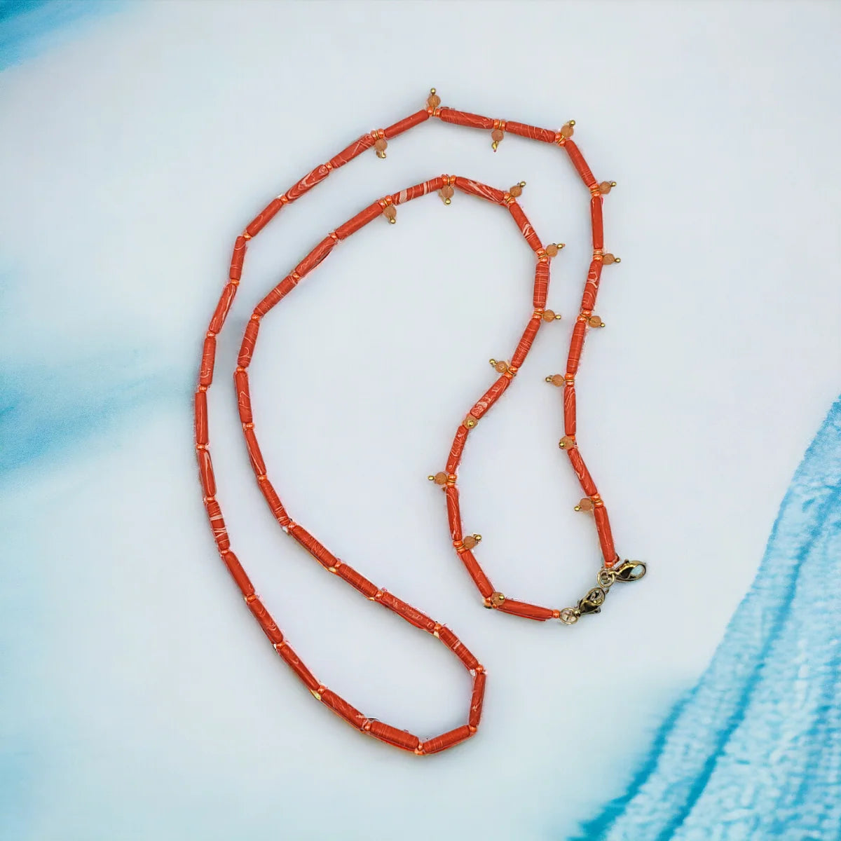 A coral-red beaded necklace with small gold-toned spacer beads and a gold clasp, arranged on a light blue and white textured background.