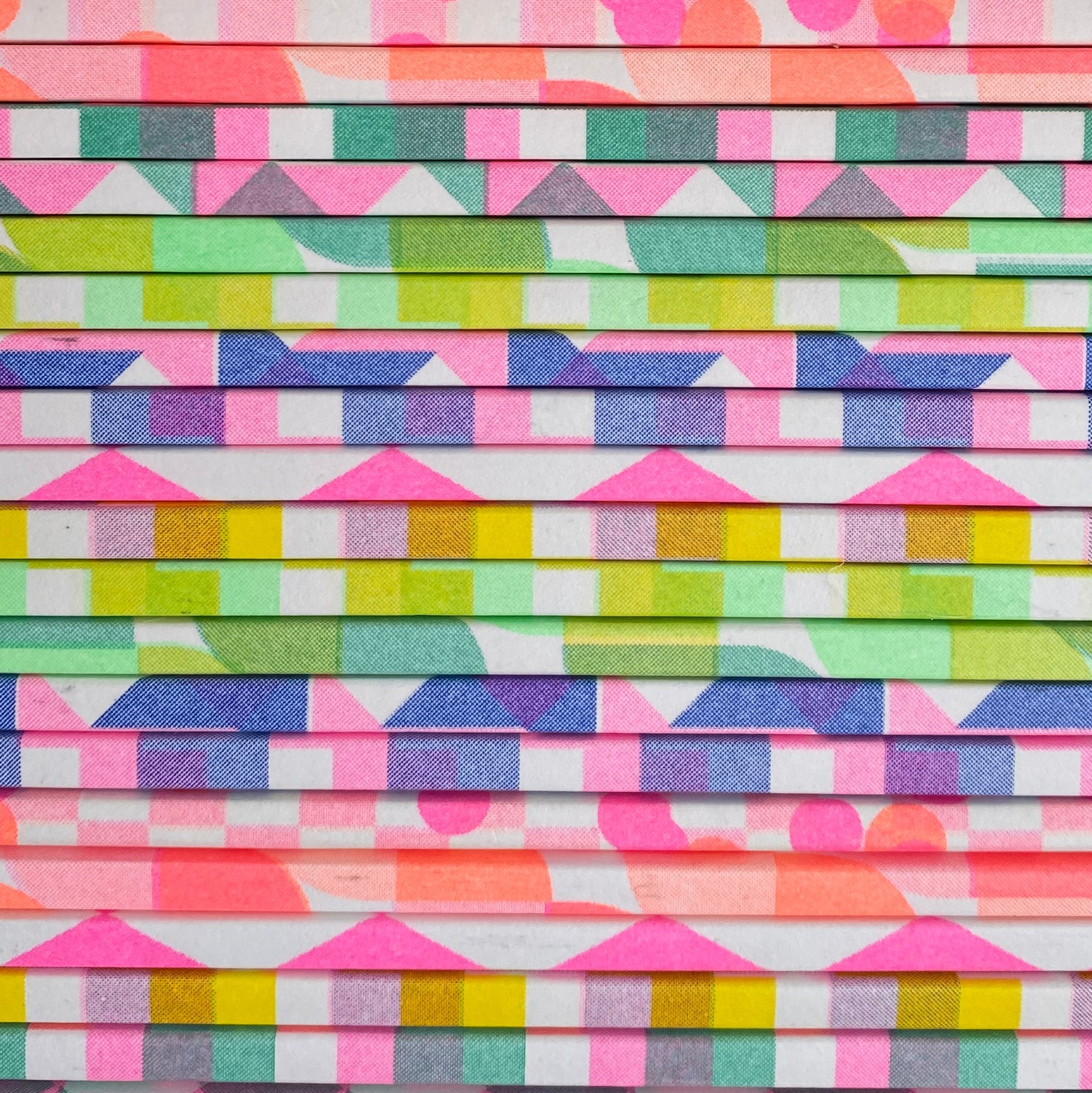 Stack of colorful papers with geometric patterns; pink, blue, green, and yellow shapes in repeating rows.