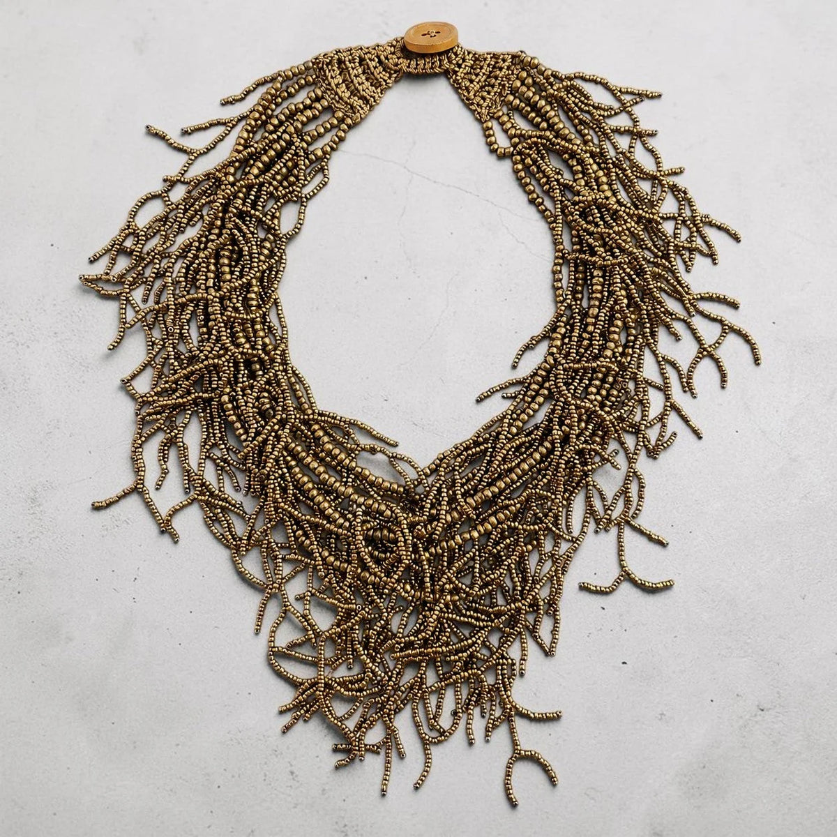A statement bib necklace made of thousands of small metallic gold beads arranged in layered fringes, featuring a single round wooden button clasp at the top, displayed on a light gray textured surface.