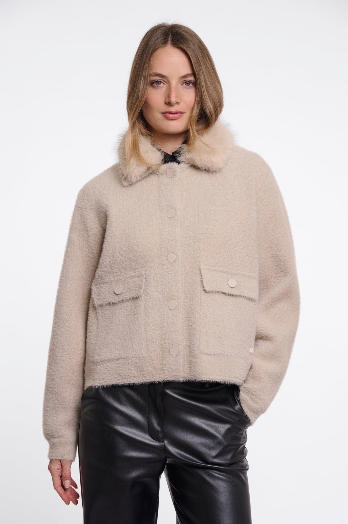 A woman wearing a beige fuzzy cropped jacket with a faux-fur collar and black leather pants, photographed against a plain light gray studio background.