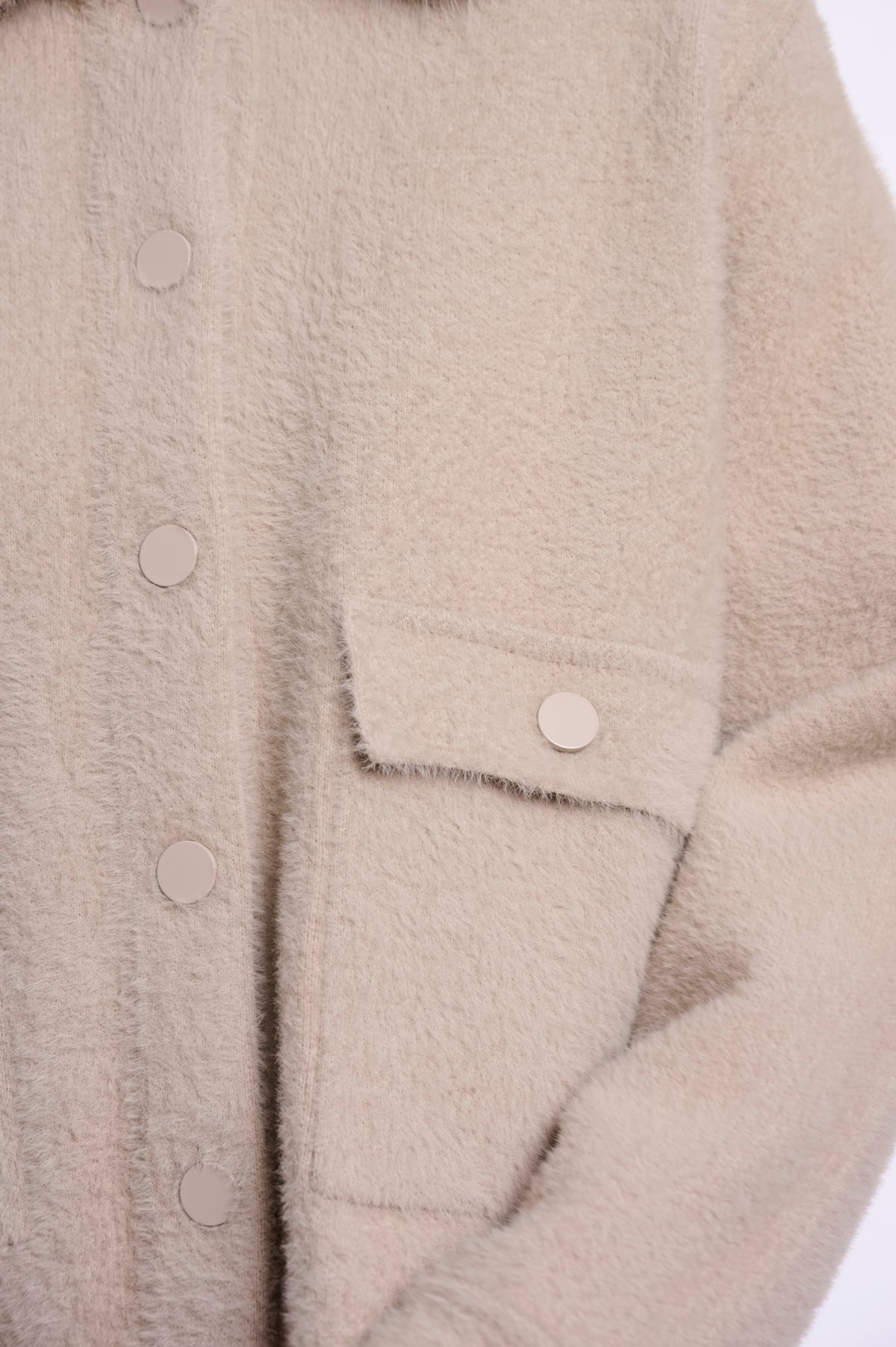 Close-up of a beige fuzzy button-front jacket or coat with a flap pocket and matching buttons, shown against a plain light background.