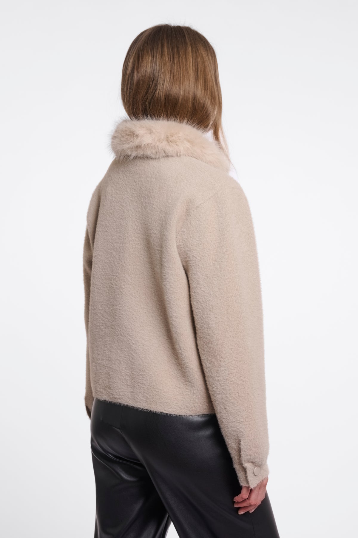 A woman shown from the back wearing a beige fuzzy cropped jacket with a fluffy collar and black pants, photographed against a clean white studio background.